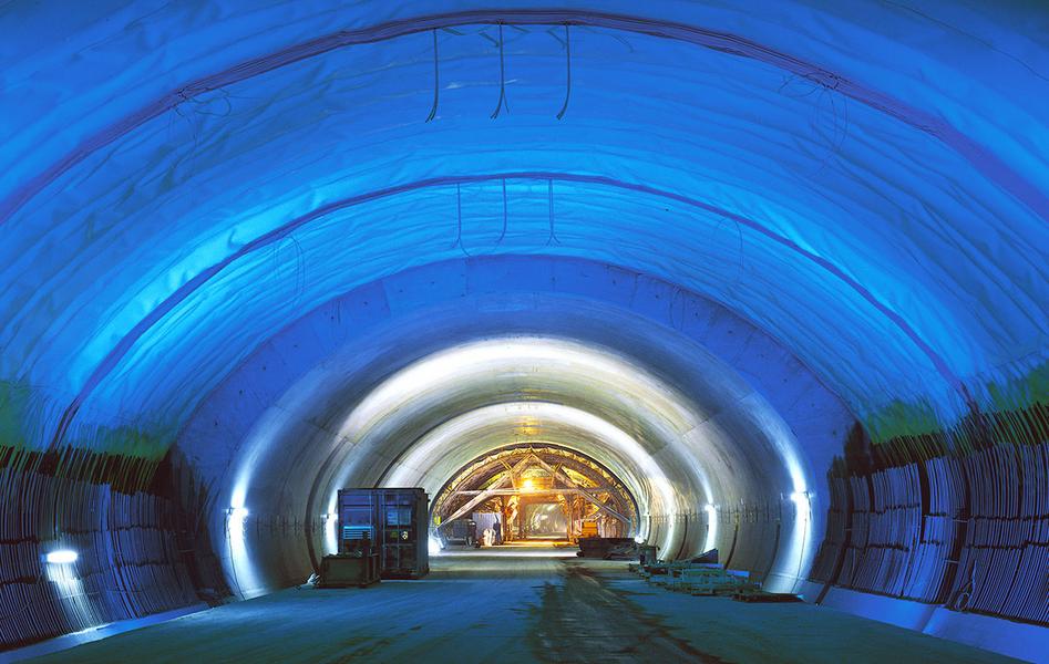 Ein beleuchteter Tunnel mit blauen Wänden und einem gewölbten Dach. Im Hintergrund sind einige Baugeräte und Personen zu sehen. Der Boden ist eben und der Tunnel scheint gut strukturiert zu sein.
