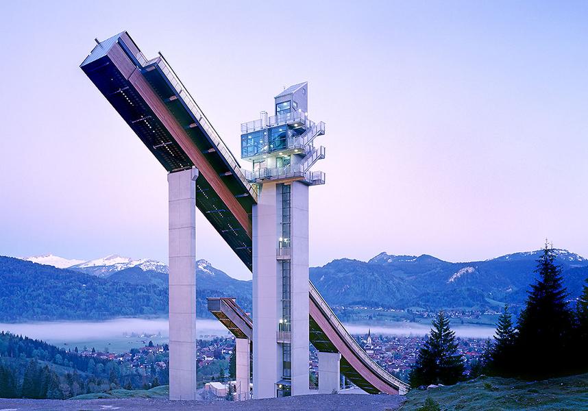 Eine Skisprungschanze steht majestätisch vor einem sanften Bergpanorama. Der hellblaue Himmel im Hintergrund verdeutlicht die Morgenstunde, während die umliegenden Wälder und das Tal leicht in Nebel gehüllt sind.
