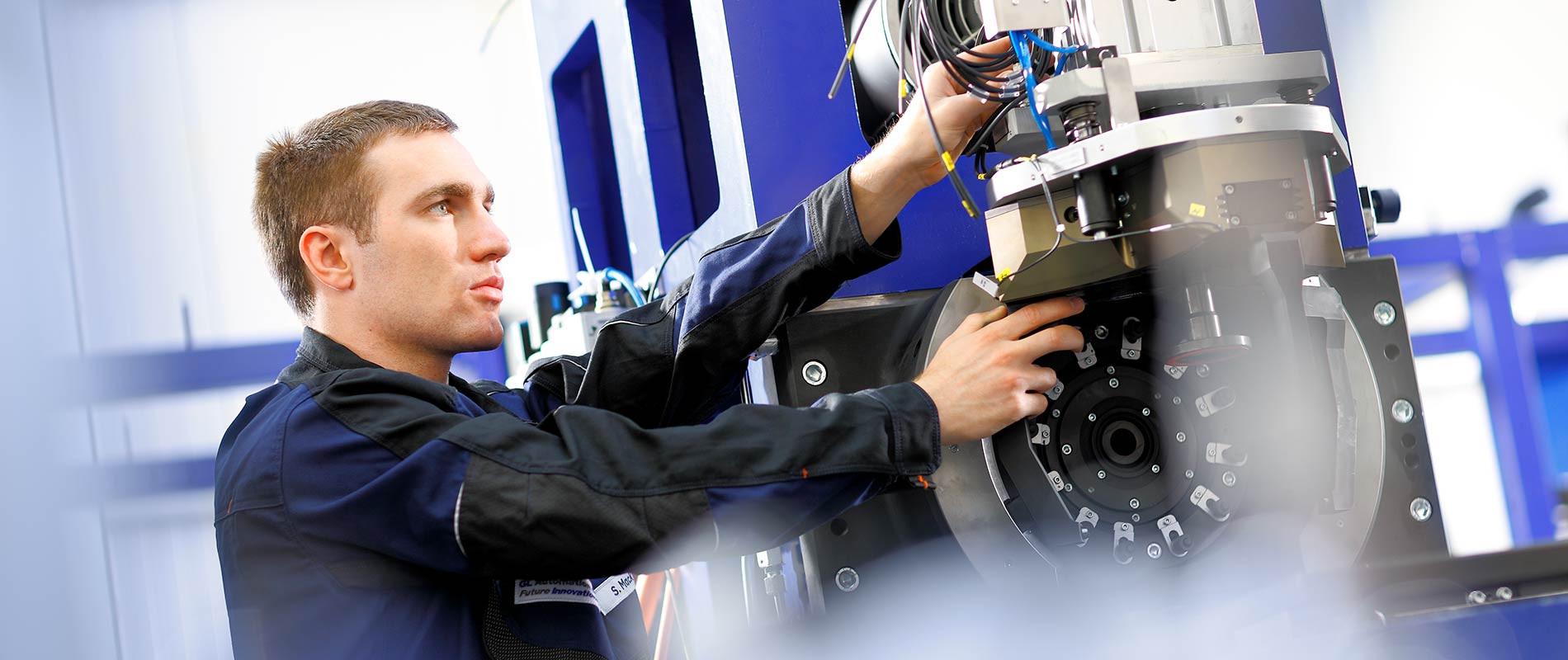 Ein Techniker arbeitet an einer Maschine, während er Kabel und technische Komponenten an einem Gerät anschließt. Er trägt eine Arbeitskleidung und konzentriert sich auf seine Aufgabe. Die Umgebung ist industriell und modern.