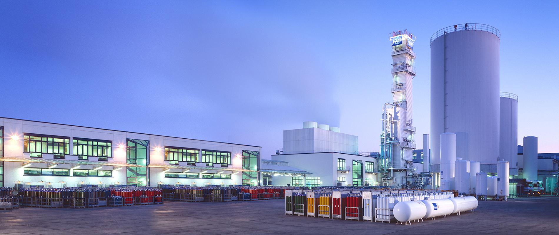 Industrieanlage bei Dämmerung mit großen Lagertanks und Gaszylindern auf einem weitläufigen Gelände. Ein hoher Turm ist im Hintergrund sichtbar, während die Gebäude in hellen Farben beleuchtet sind.