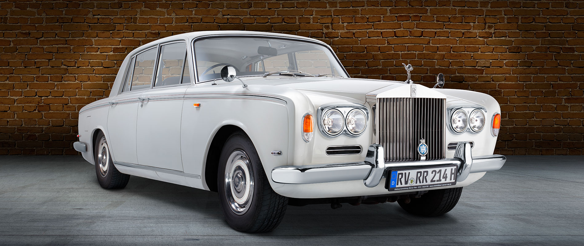 Ein eleganter, weißer Rolls-Royce Limousine steht vor einer Backsteinmauer. Der Wagen hat klassische Linien und eine markante Kühlerfigur. Die glänzenden Scheinwerfer und Felgen verleihen dem Auto einen luxuriösen Look.